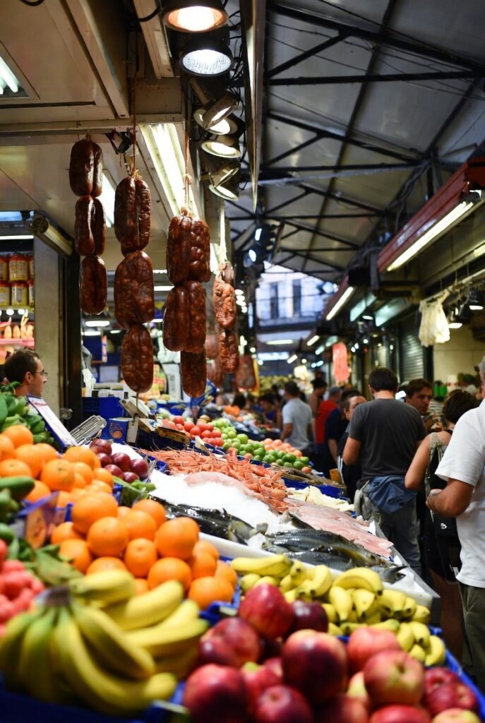 vibrante do Mercado La Boquería em Barcelona com frutas coloridas, jamón ibérico pendurado, frutos do mar frescos no gelo, vendedores e clientes. Iluminação natural quente, estilo fotográfico de viagem gastronômica.