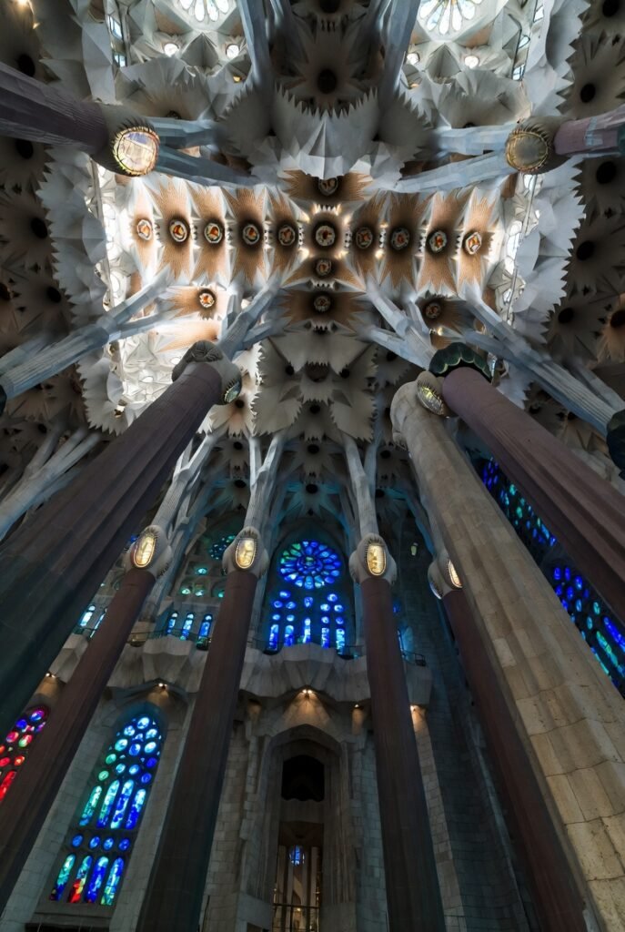 Experiencias imperdiveis em barcelona. Vista para cima do interior impressionante da Sagrada Família em Barcelona, com colunas ramificadas como árvores, vitrais coloridos em azul e vermelho iluminando a nave. Ultra grande angular, fotografia cinematográfica.